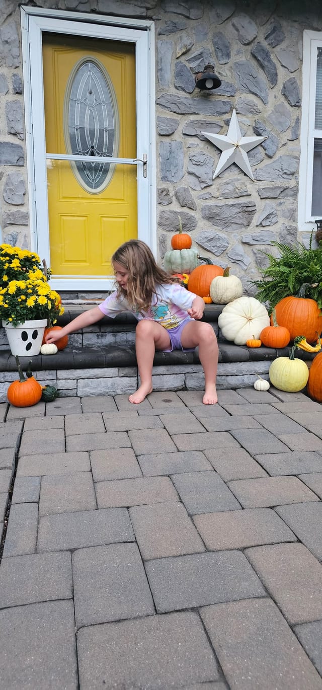 A girl on stone steps reaching for a small white pumpkin, beside a yellow front door framed with pumpkins and mums